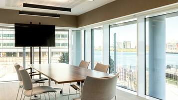 Un salón de conferencias con una gran mesa, sillas, un televisor de pantalla plana y ventanas amplias que dan a una masa de agua y edificios.