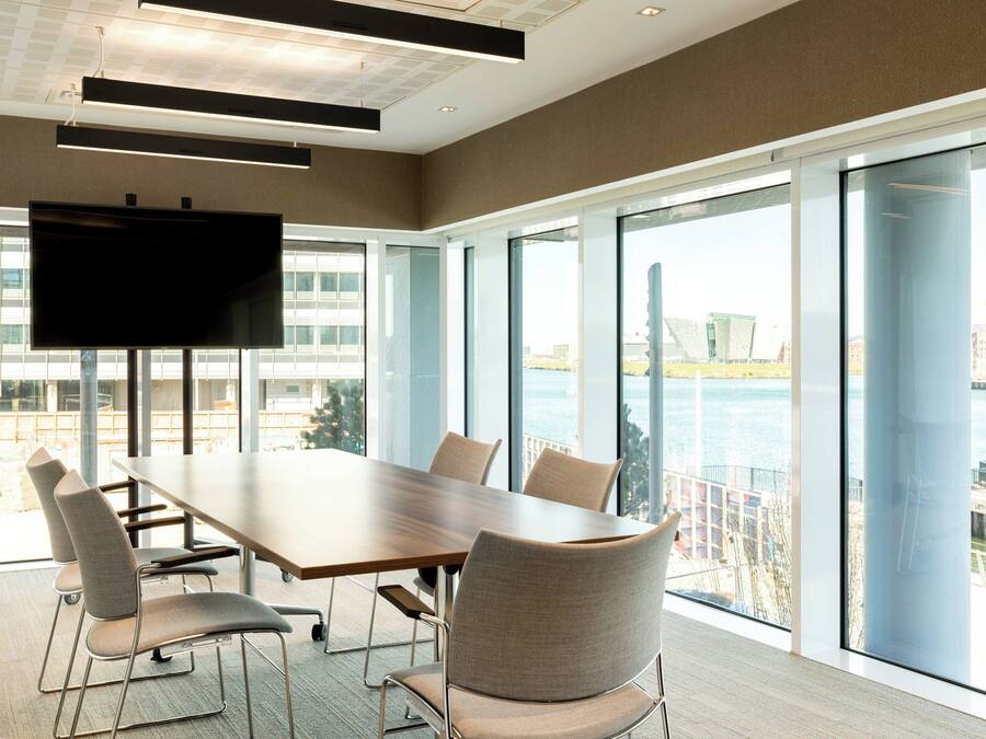 Un salón de conferencias con una gran mesa, sillas, un televisor de pantalla plana y ventanas amplias que dan a una masa de agua y edificios.