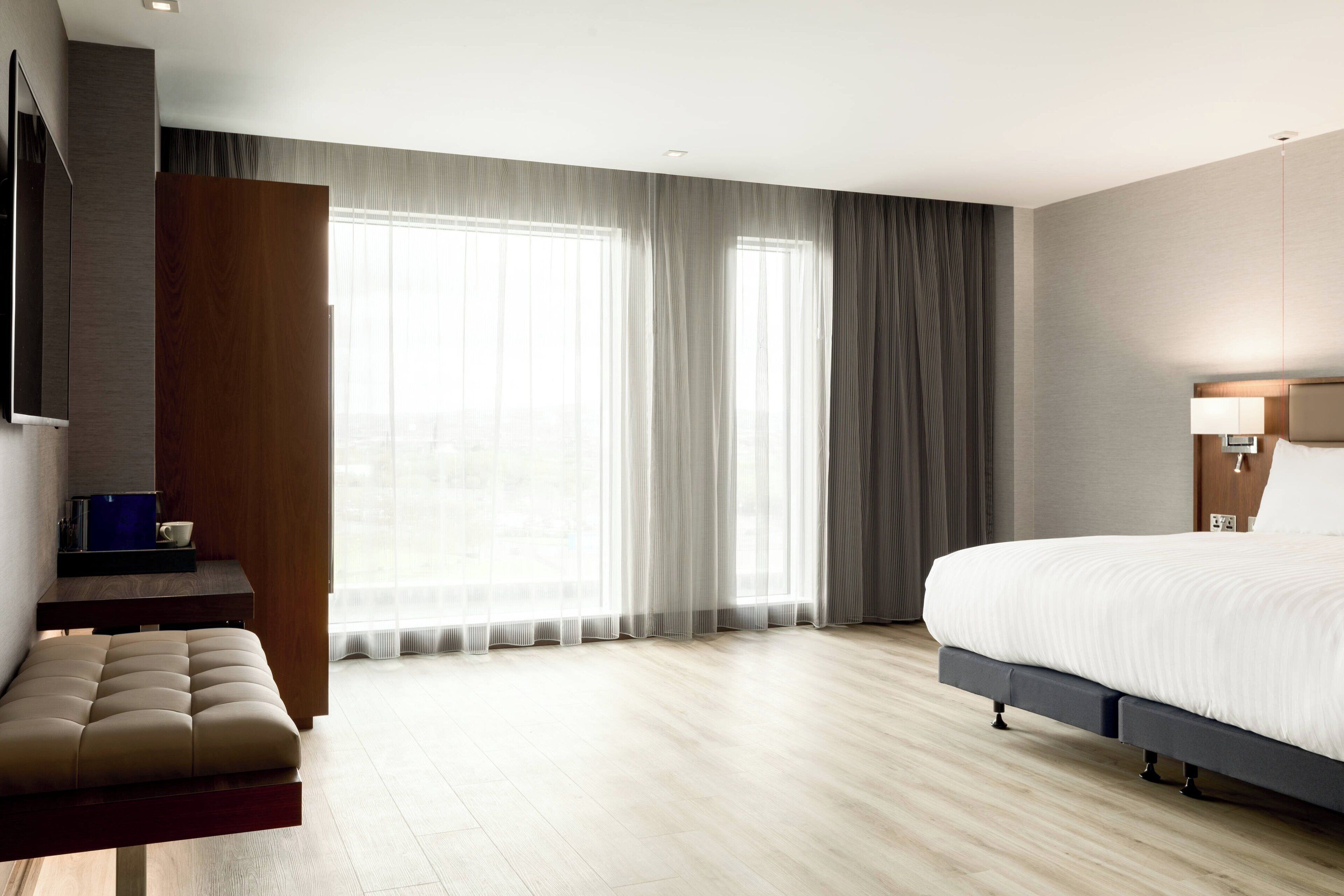 Una habitación de hotel moderna con una cama grande, un banco de madera, un televisor y ventanas amplias con cortinas translúcidas.