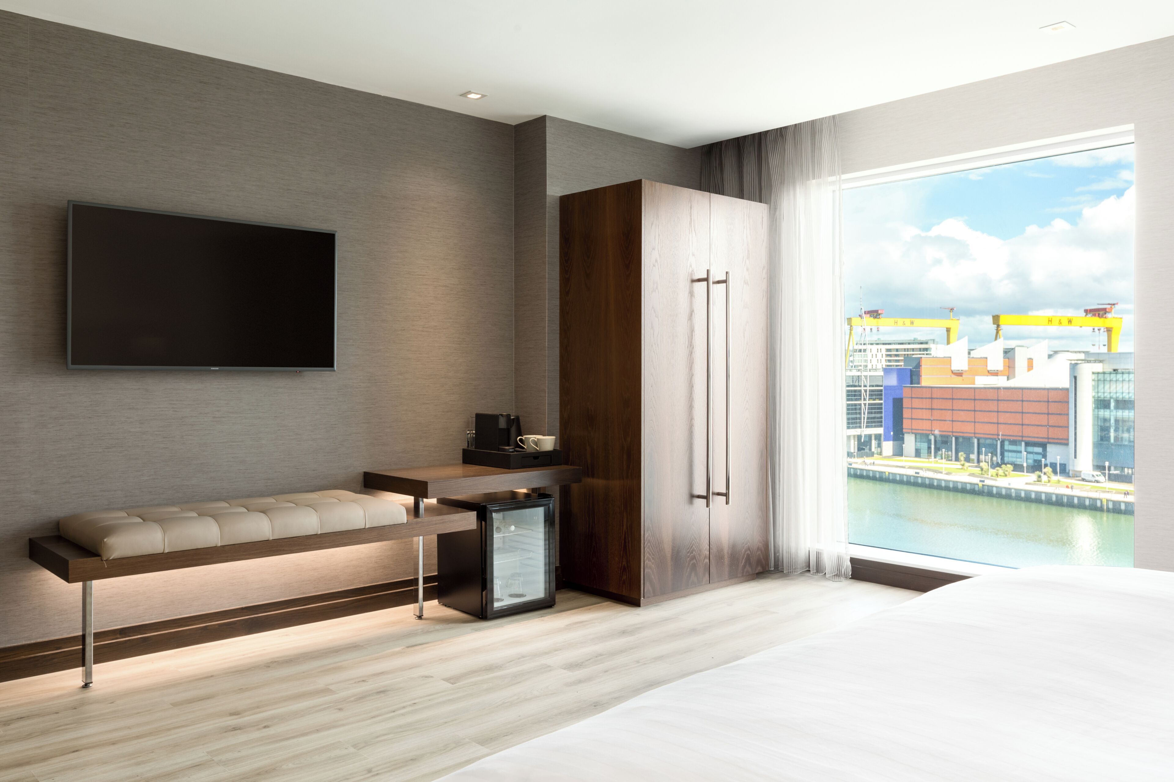 Une chambre d’hôtel moderne dotée d’une télévision à écran plat, d’un canapé, d’un mini-réfrigérateur et d’une grande fenêtre donnant sur la ville.