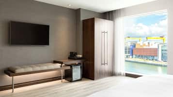 Une chambre d’hôtel moderne dotée d’une télévision à écran plat, d’un canapé, d’un mini-réfrigérateur et d’une grande fenêtre donnant sur la ville.
