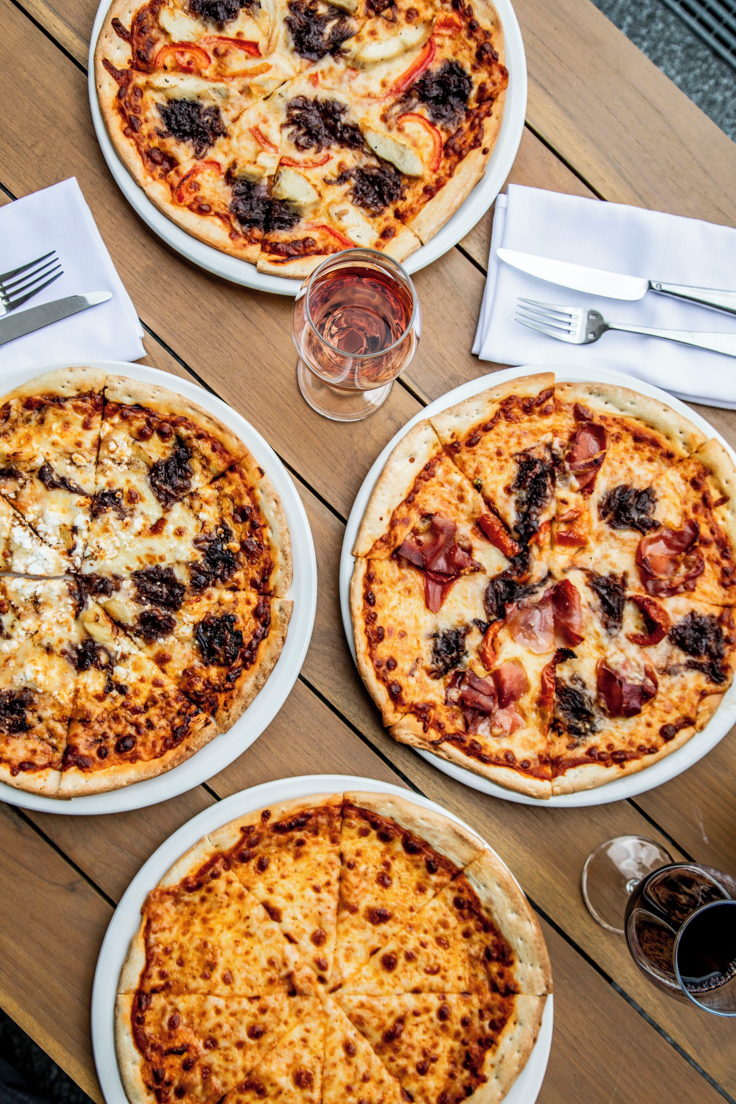 A wooden table with four pizzas, each with different toppings, and a glass of red wine.