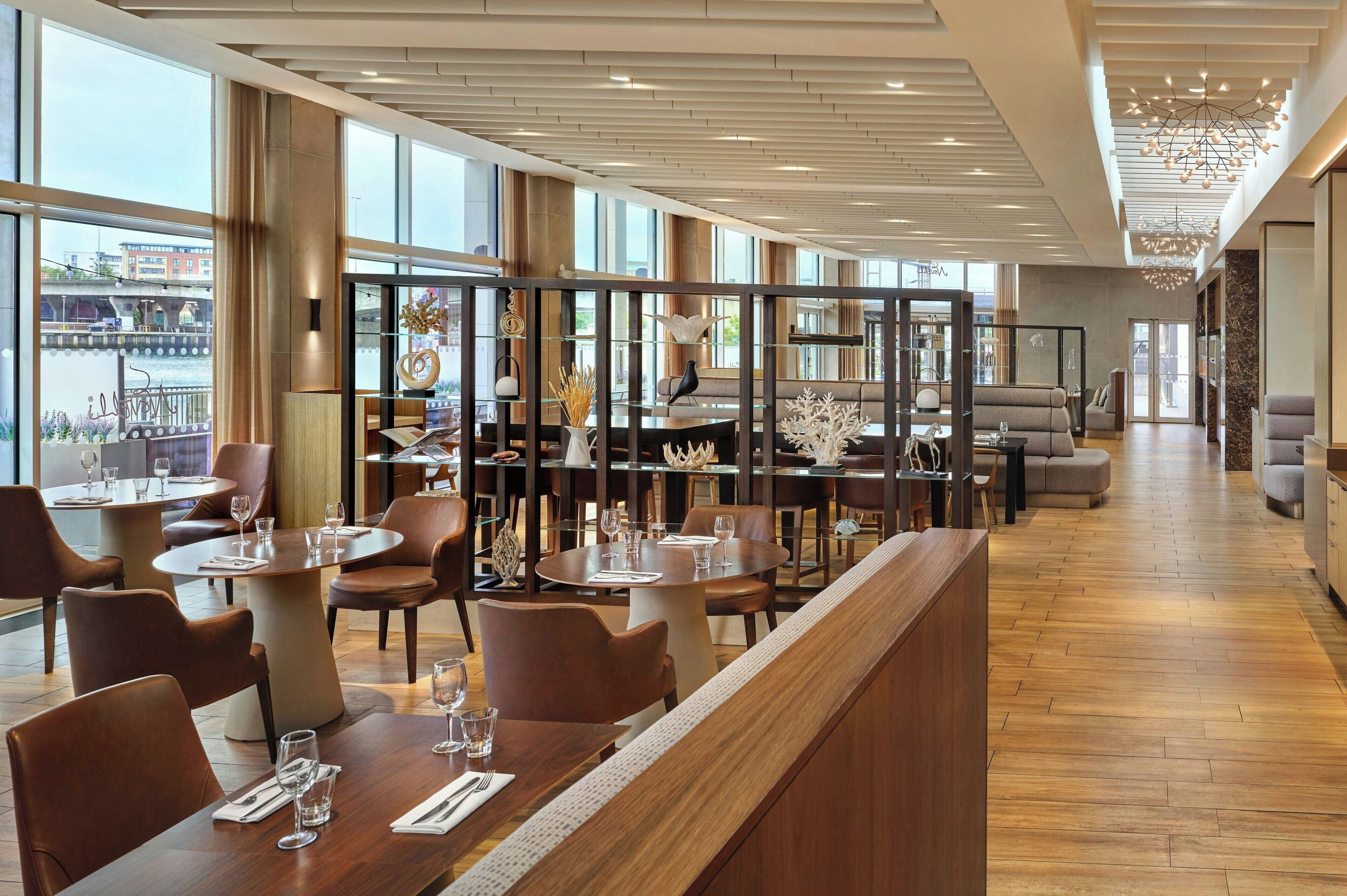 A modern restaurant with high ceilings, large windows, and a variety of seating options including round tables and sofas.