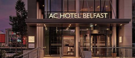 The exterior of AC Hotel Belfast with a modern facade, large glass entrance, and a staircase leading to the entrance.