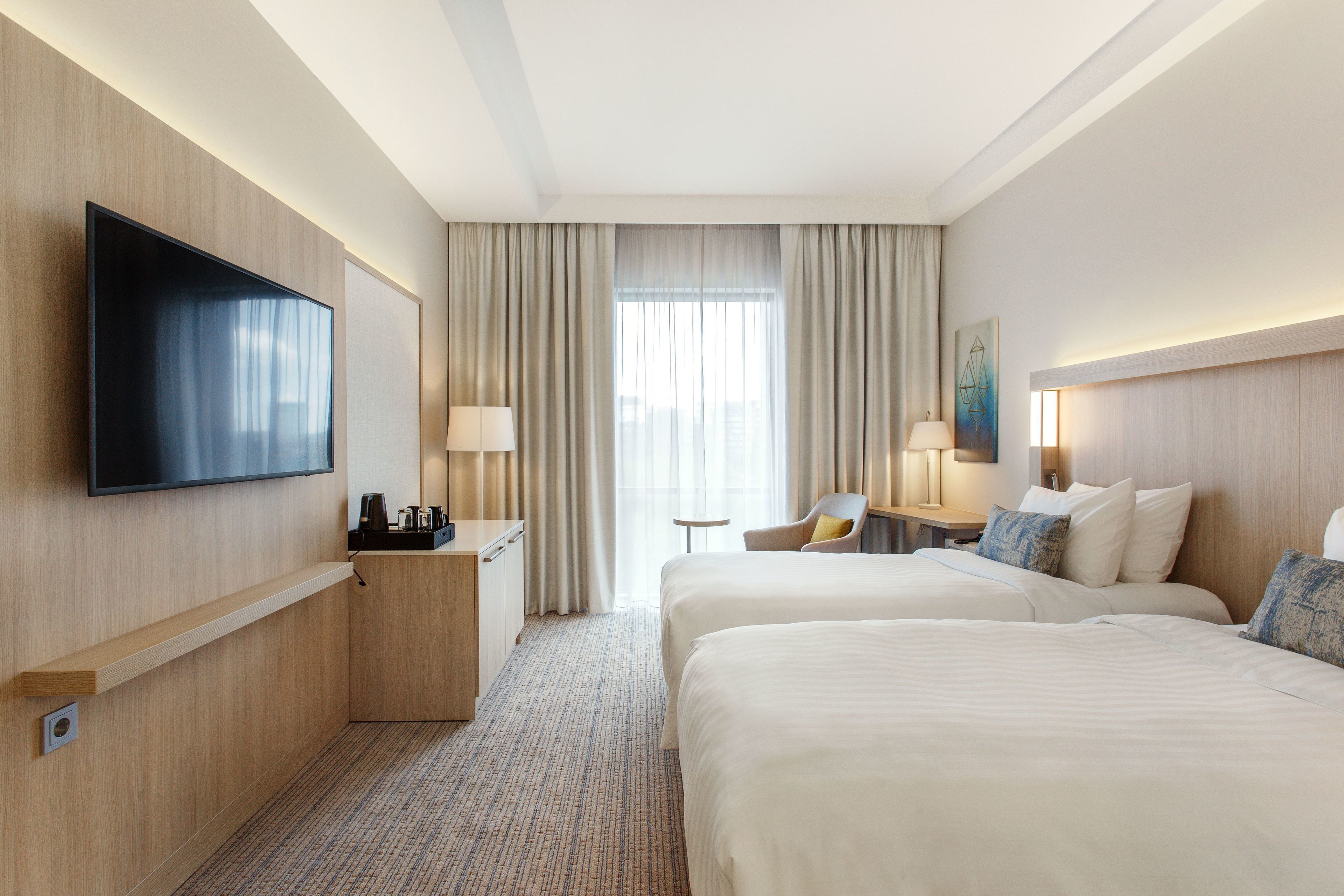 Habitación de hotel con dos camas, un televisor de pantalla plana, un escritorio con lámpara y vistas a la ciudad a través de amplias ventanas.
