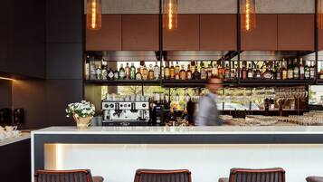Un espace bar moderne comprenant un comptoir, trois tabourets, des luminaires suspendus et une variété de bouteilles et de verres disposés sur des étagères.