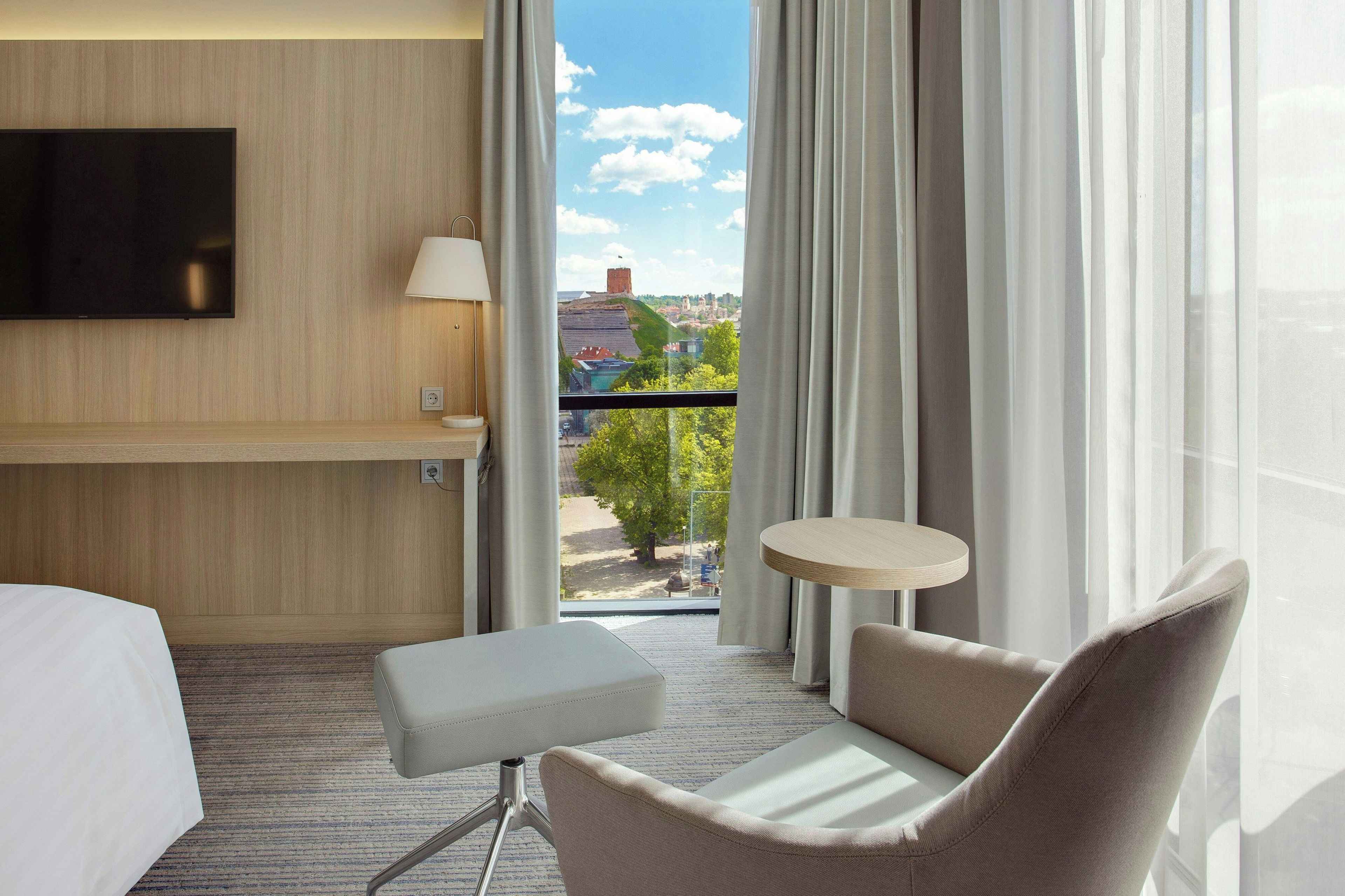 A hotel room with a bed, a TV mounted on the wall, a small table, a chair, and a view of a cityscape through the window.