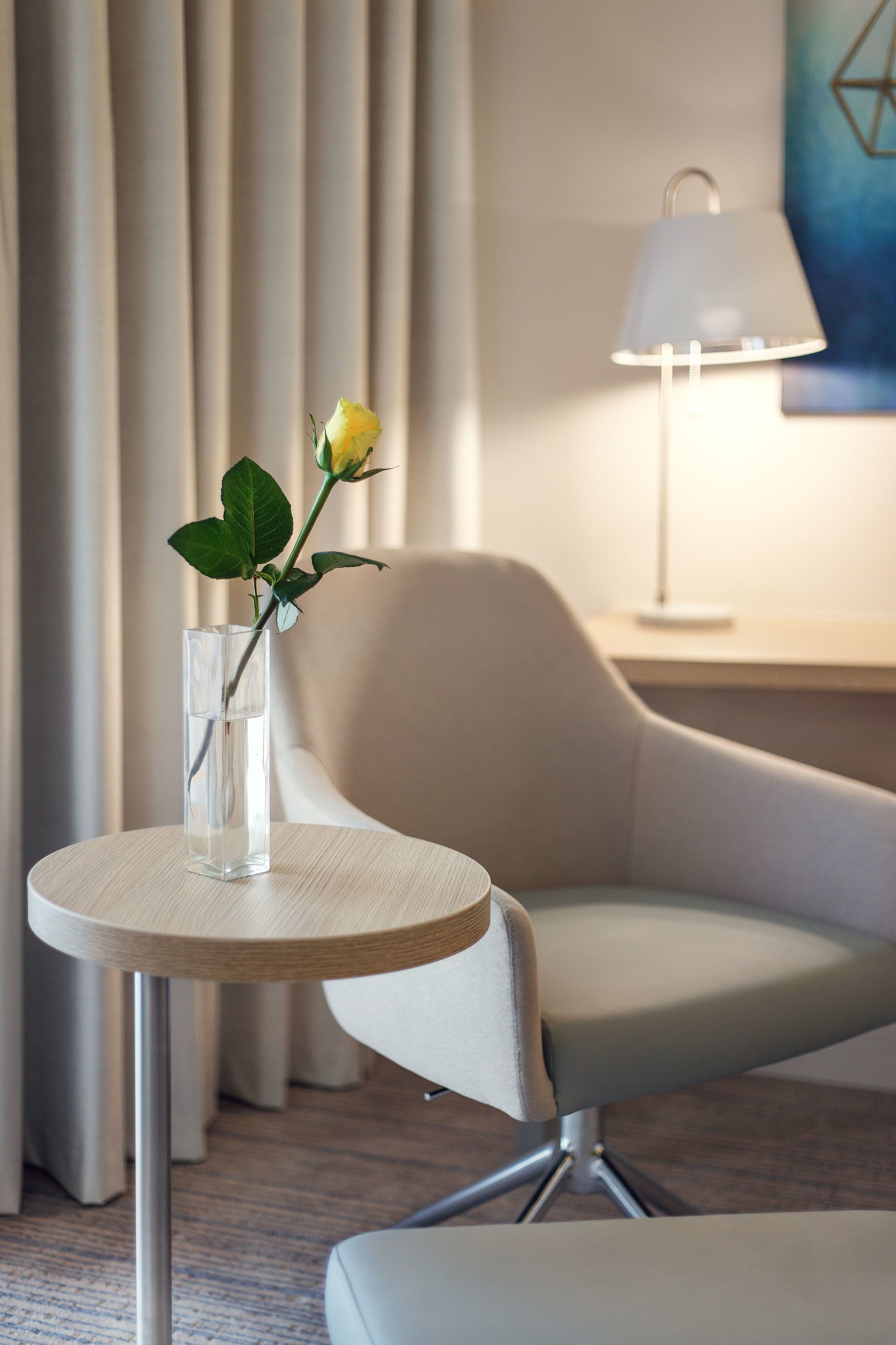 A modern hotel room with a round table, a chair, a vase with a single yellow flower, and a lamp on a bedside table.
