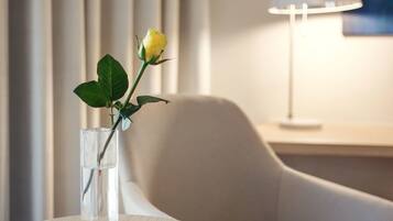 A modern hotel room with a round table, a chair, a vase with a single yellow flower, and a lamp on a bedside table.