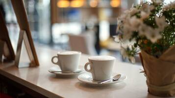 Dos tazas blancas sobre platillos en una mesa, con una cuchara, un florero y un fondo desenfocado que sugiere un café.