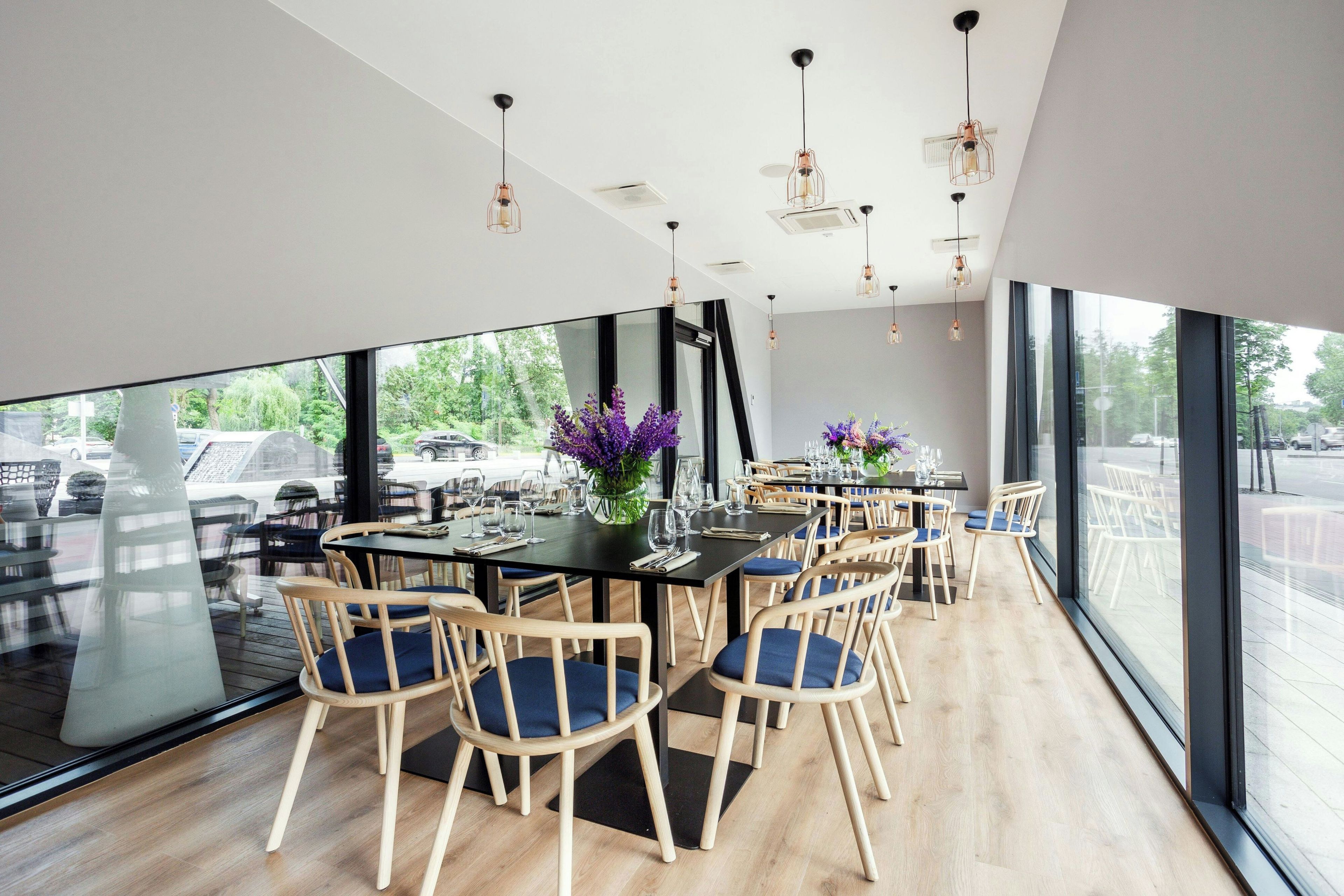 Un comedor moderno con sillas y mesas de madera, ventanas amplias y lámparas colgantes.