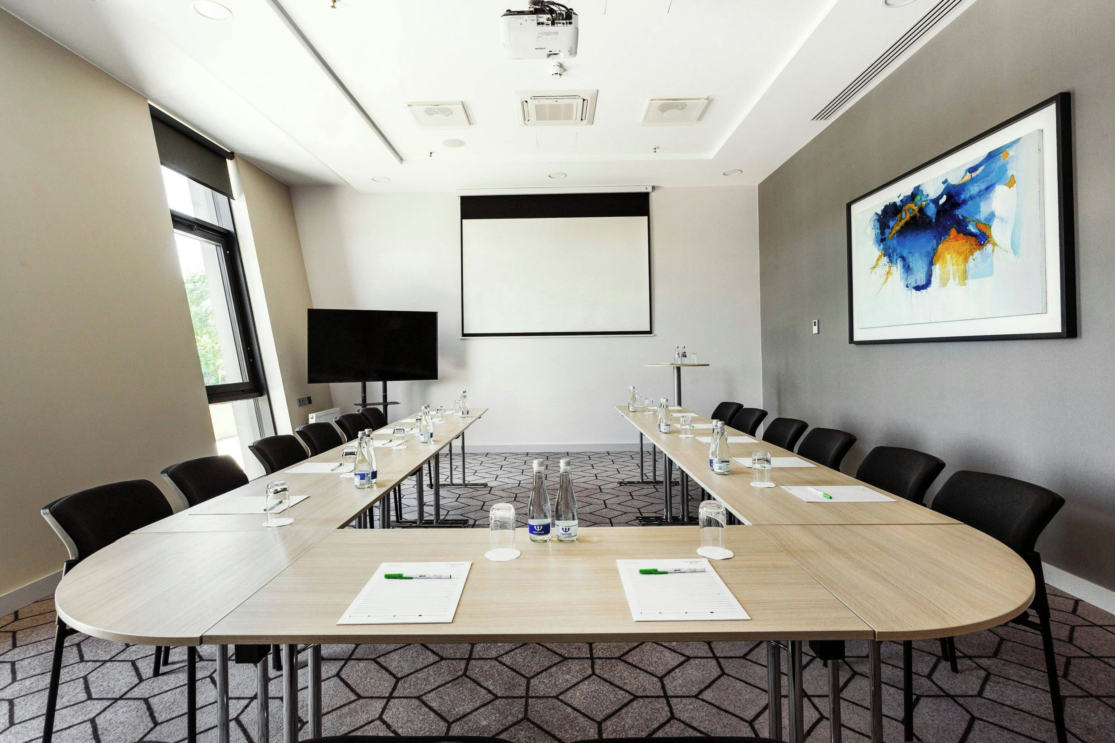 Un salón de conferencias con una larga mesa, sillas, una pantalla de proyector y un cuadro abstracto grande en la pared.