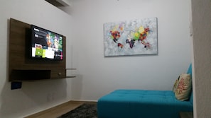 Una sala de estar con una televisión de pantalla plana montada en un estante de madera, un cuadro abstracto de colores en la pared y un sofá turquesa con un cojín decorativo.