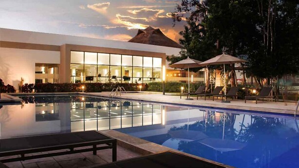 A hotel pool area with lounge chairs, a glass-enclosed building, and a mountain view. - Tu Casa en Playa del Carmen conSportClub (Playa del Carmen)
