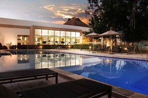 Un espace piscine d’hôtel avec des chaises longues, un bâtiment entièrement vitré et une vue sur les montagnes.