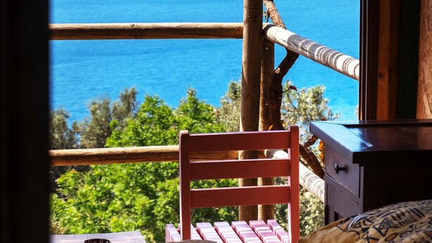 A room with a wooden bed, a small table, and a chair. A window offers a view of the sea and mountains. - Deep Ocean Camping - Adults Only (Fethiye)