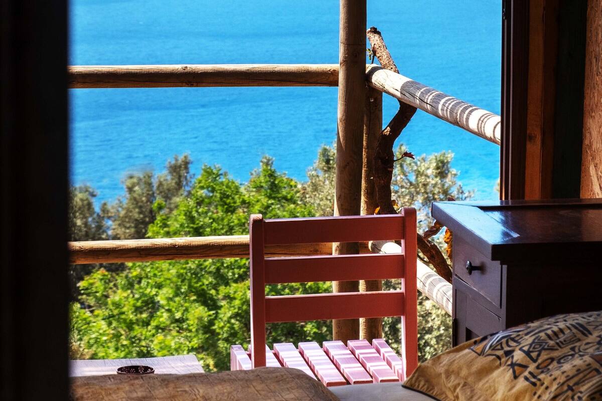 A room with a wooden bed, a small table, and a chair. A window offers a view of the sea and mountains.
