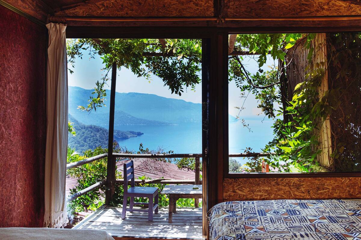 A room with a large window offering a view of a mountainous landscape and a body of water. There is a wooden deck outside with a table and chairs.