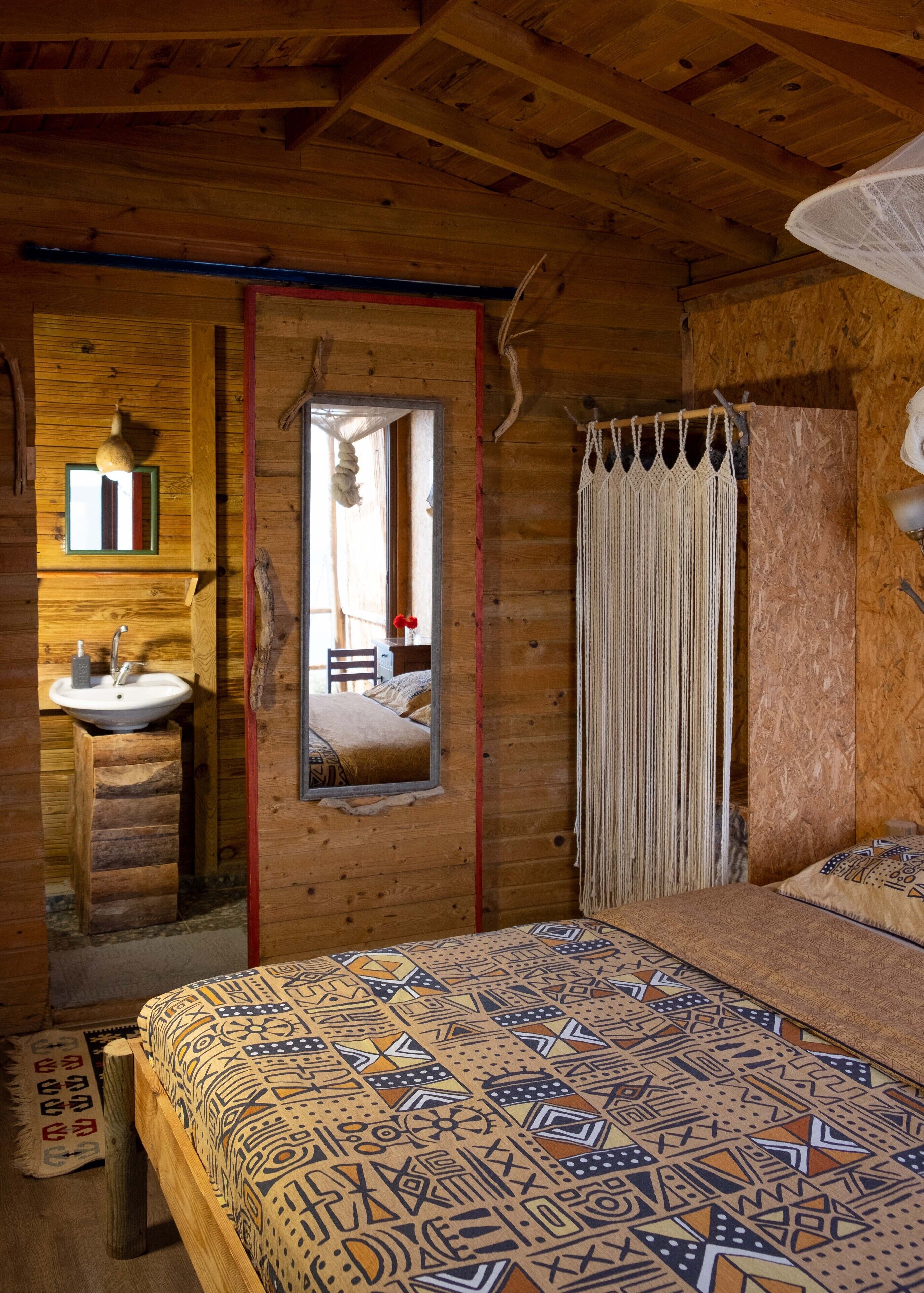 Un dormitorio de cabaña de madera con cama, espejo, lámpara colgante y cortina de macramé.