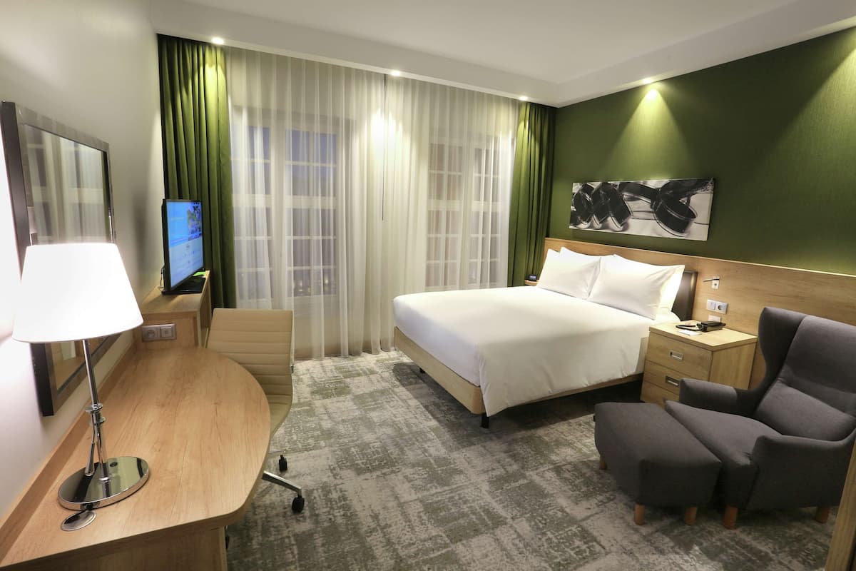 Ein Hotelzimmer mit Bett, Schreibtisch mit Lampe, Stuhl, Fernseher und einer grünen Wand mit einem Gemälde.