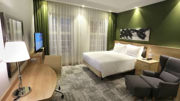 A hotel room with a bed, a desk with a lamp, a chair, a TV, and a green wall with a painting.