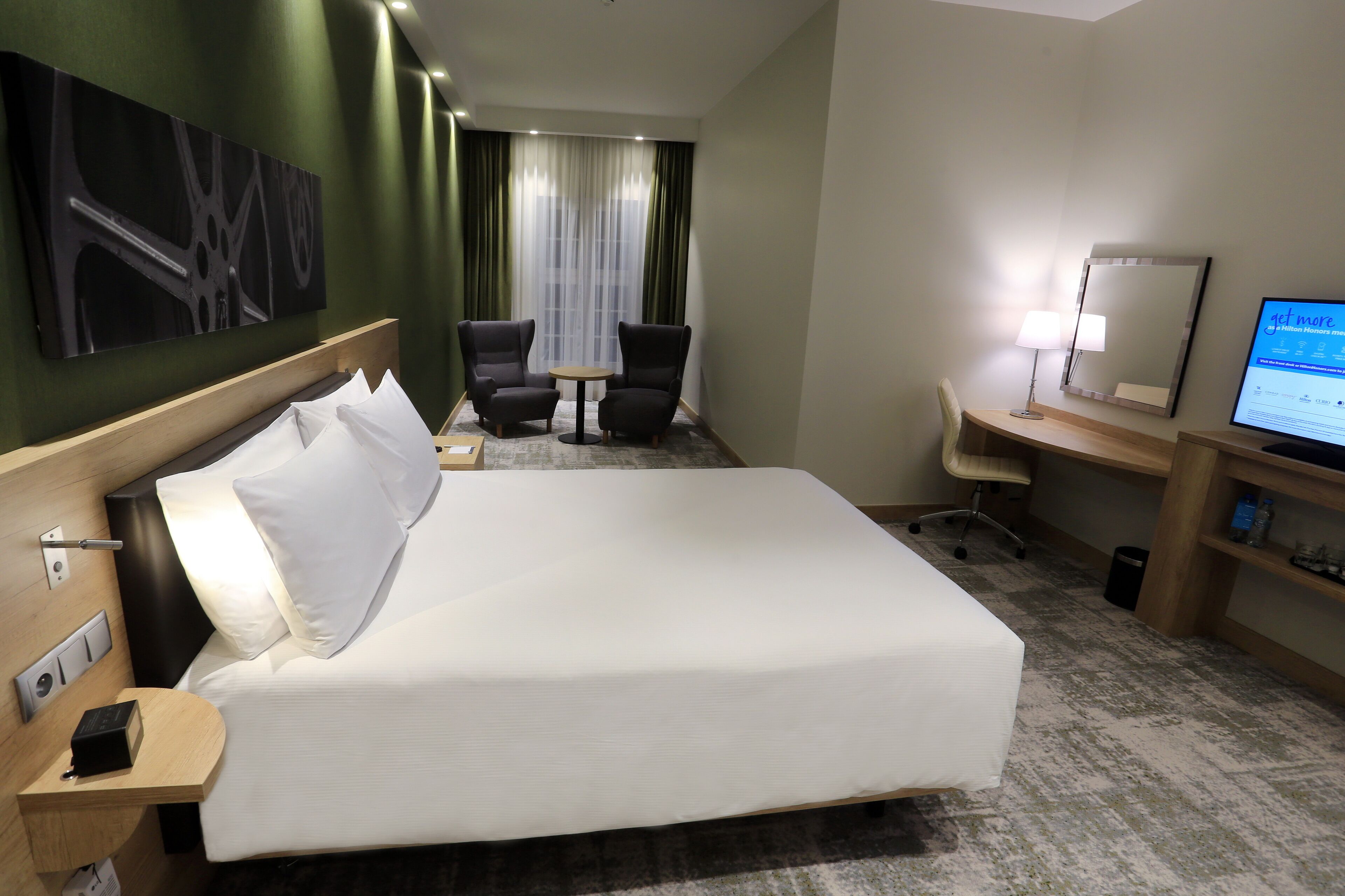 Habitación de hotel con una cama grande, un escritorio con computadora, dos sillas, una mesita y un ventanal con cortinas.