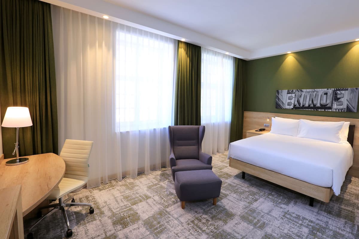Ein Hotelzimmer mit Bett, Schreibtisch mit Lampe, Stuhl und großem Fenster mit Vorhängen.