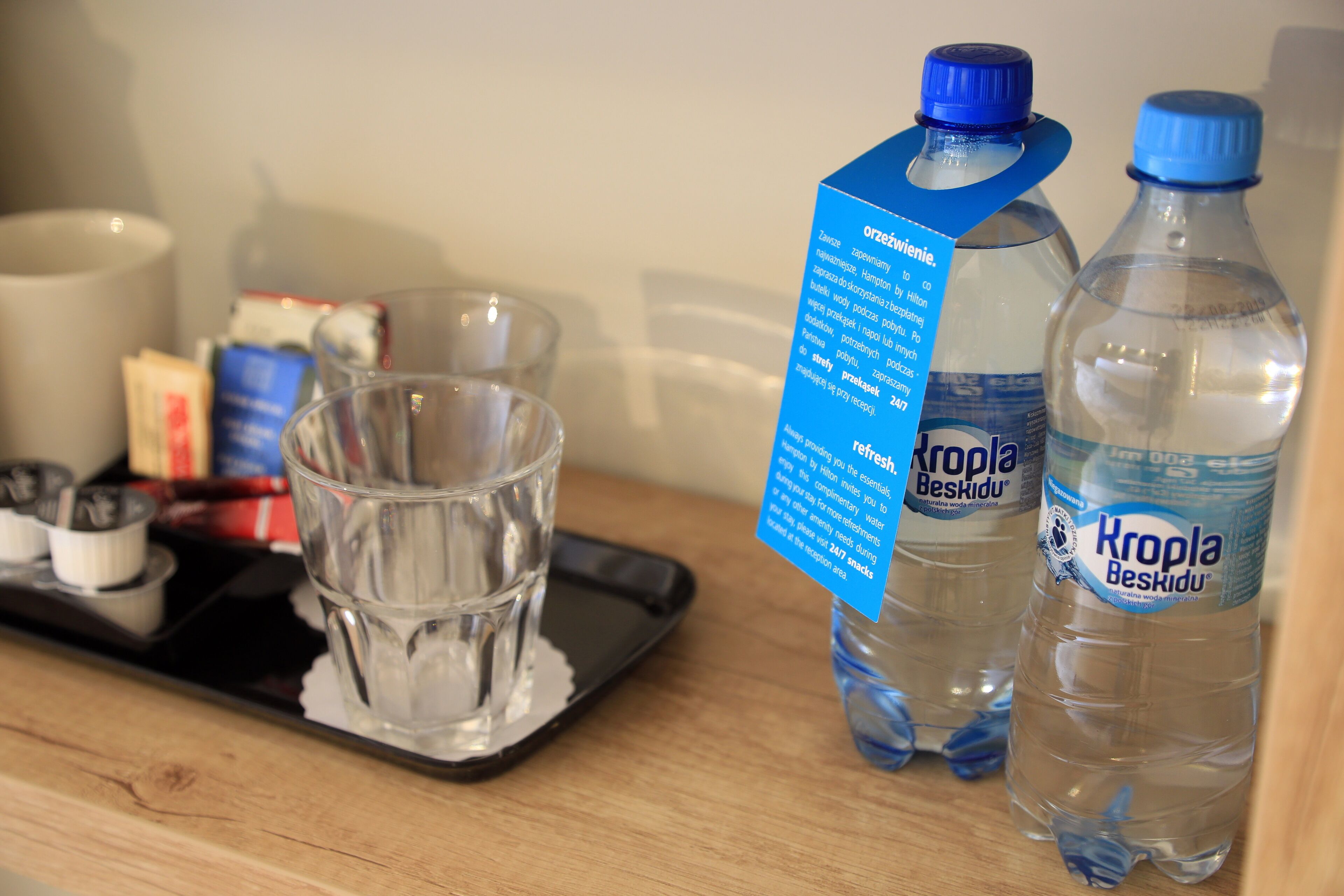 Een dienblad met een glas water, twee flessen Kropla Beskidu water, en een blauw informatief kaartje.