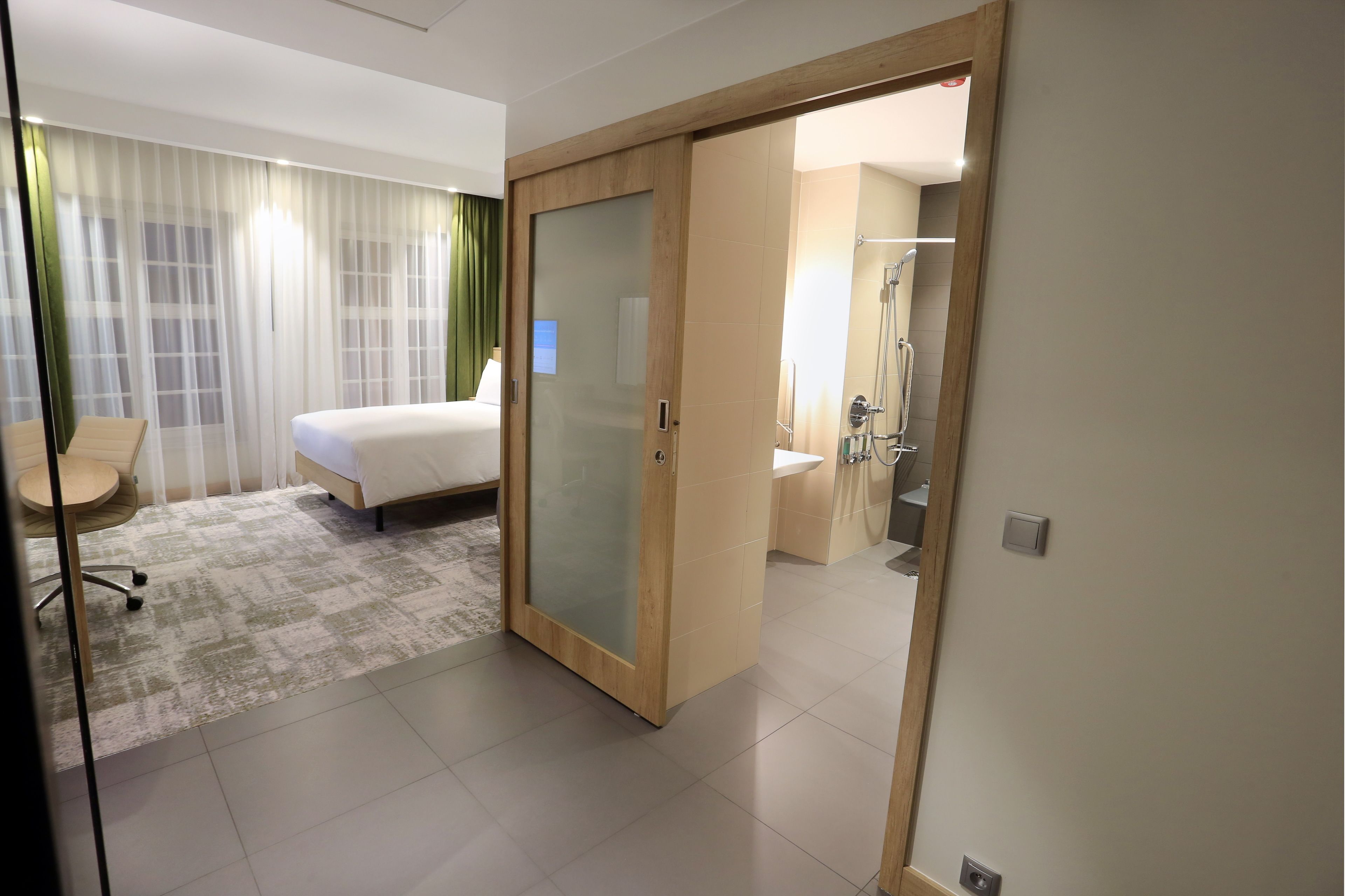 A hotel room with a bed, a desk, a chair, a bathroom with a shower, and a glass door leading to another room.