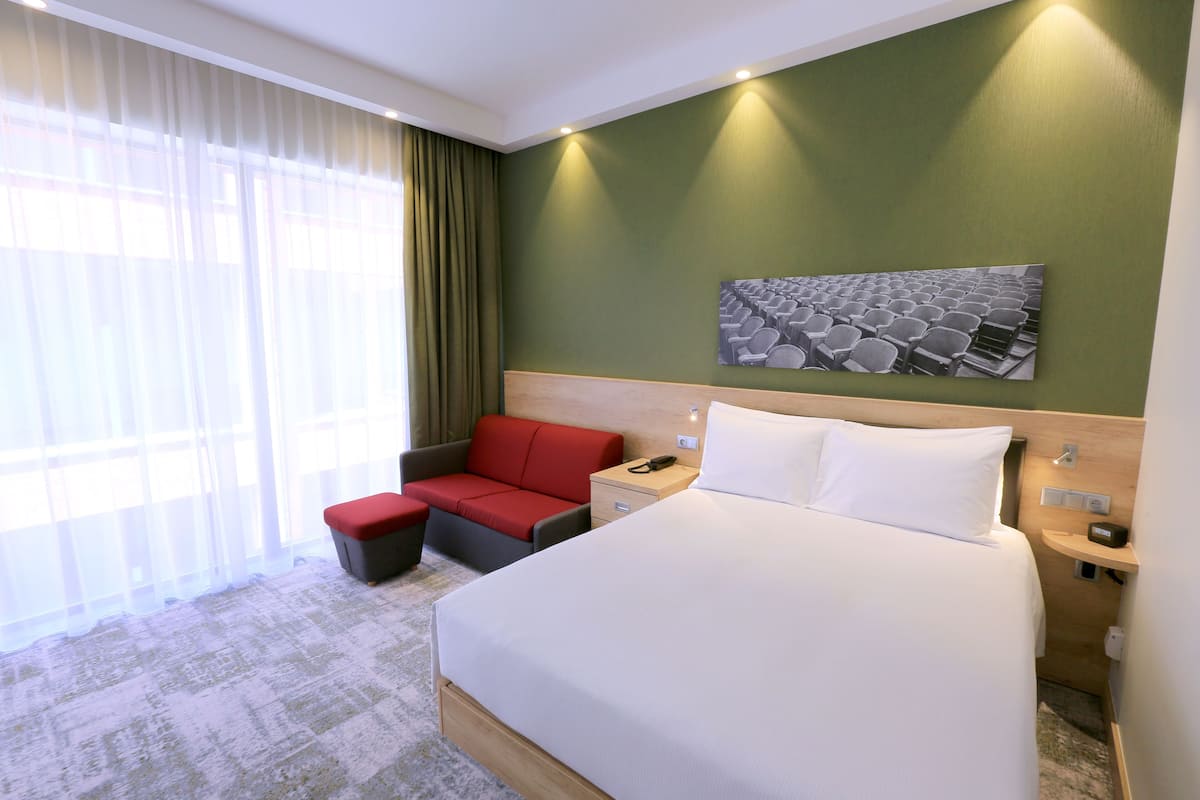 Ein Hotelzimmer mit einem großen Bett, einem roten Schlafsofa, einem Nachttisch und einem Wandbild.