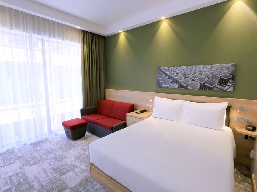 A hotel room with a large bed, a red sofa, a small table, and a wall-mounted artwork.