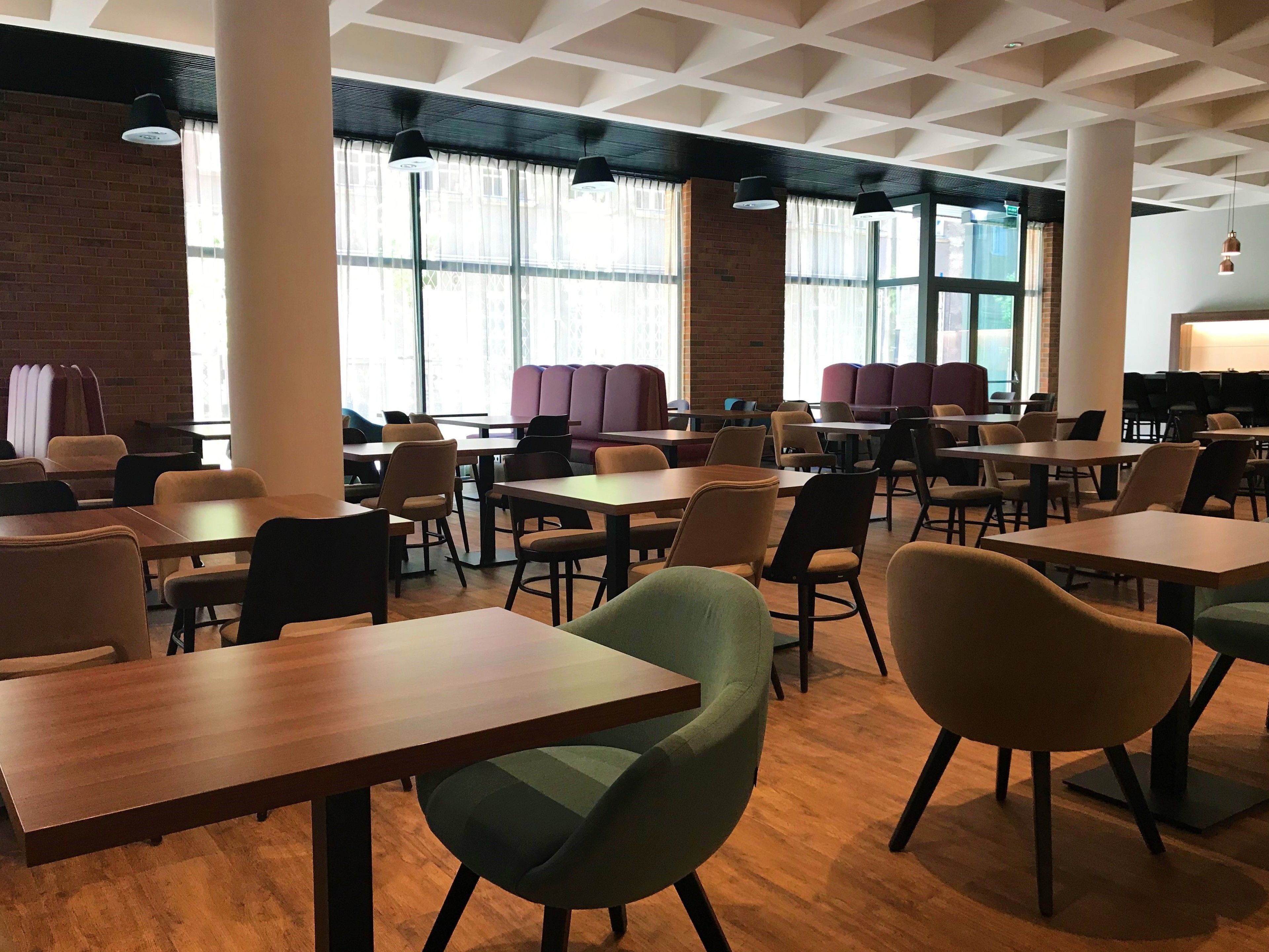 A modern dining area with wooden tables and chairs, large windows with curtains, and a brick wall.