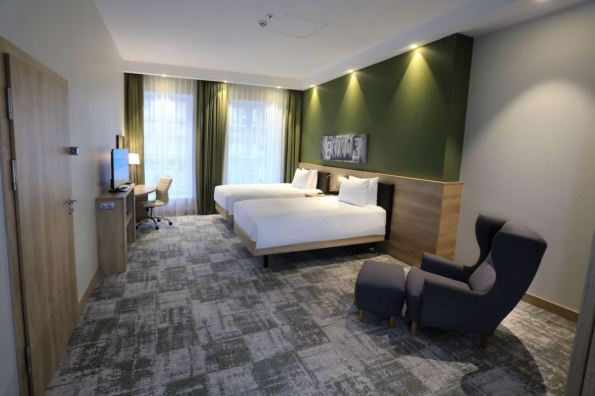 Ein Hotelzimmer mit zwei Betten, einem Schreibtisch mit Computer, einem Stuhl und einem großen Fenster mit Vorhängen.