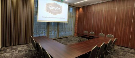 A conference room with a long wooden table, six chairs, a projection screen displaying 'Hampton by Hilton Gdansk Old Town', and a view of a snowy landscape through large windows.