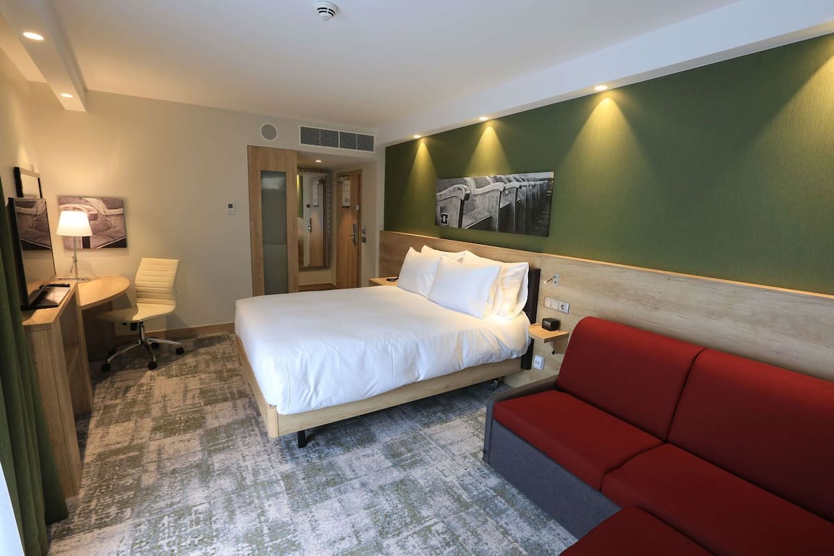 Ein Hotelzimmer mit einem Bett, einem Schreibtisch, einem Sessel und einem roten Sofa. An der Wand hängt ein Gemälde.