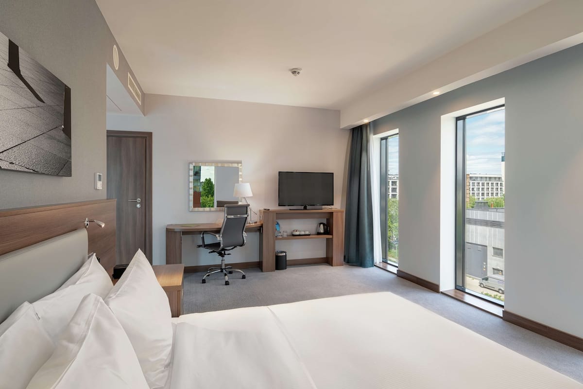 Ett hotellrum med en stor säng, ett skrivbord med en dator, en tv och utsikt över staden genom stora fönster.