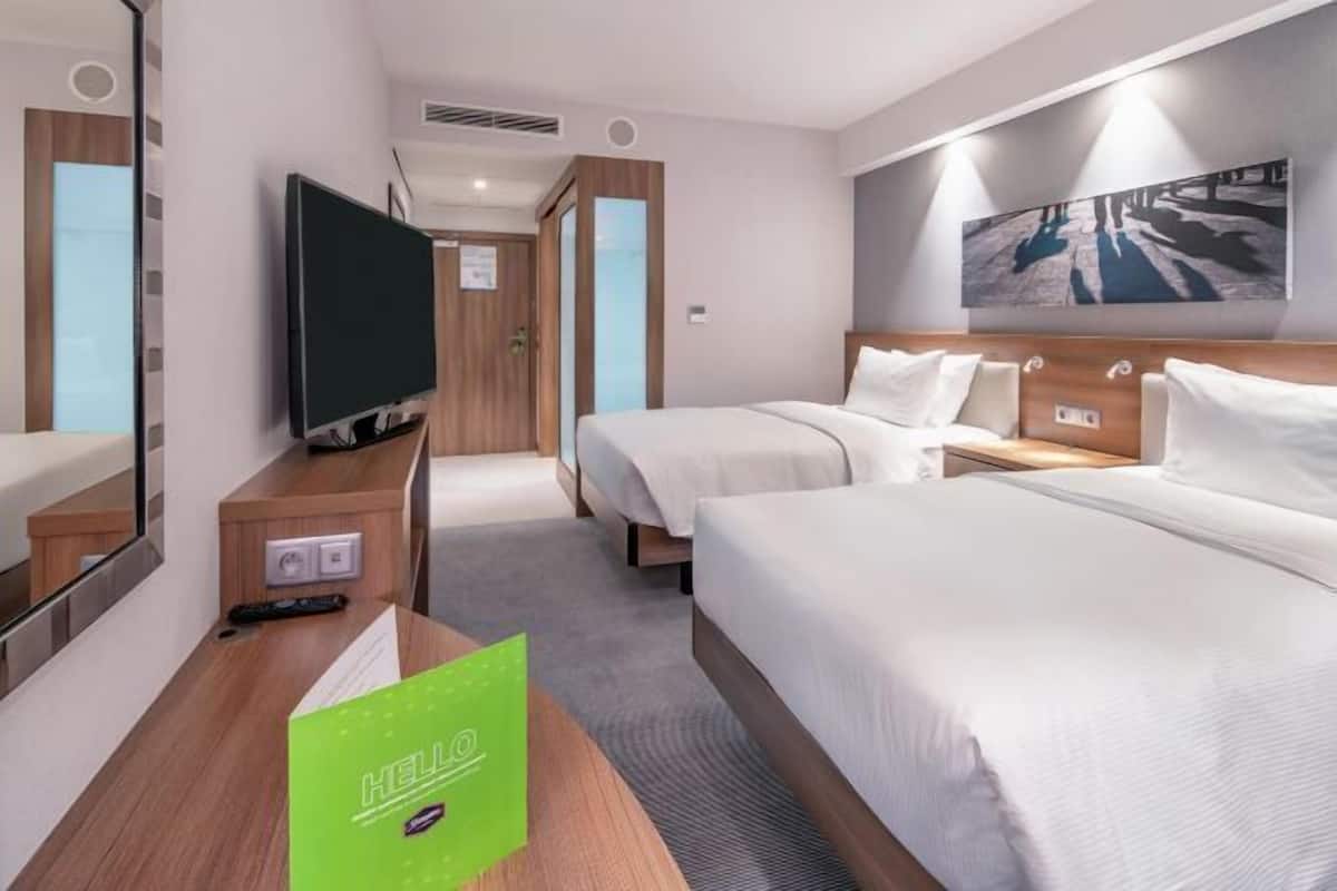 Ett hotellrum med två sängar, en tv, en spegel och en tavla på väggen.