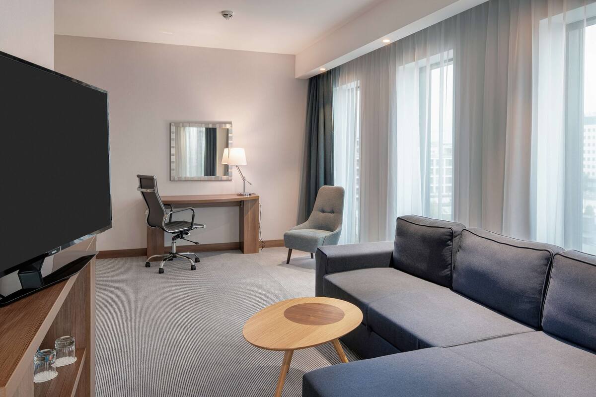 Ett modernt hotellrum med en stor platt-TV, en soffa, en fåtölj, ett skrivbord med en lampa och ett stort fönster med gardiner.