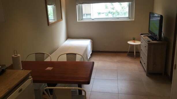 Une petite pièce bien éclairée, avec un lit, un bureau, une chaise, une télévision et une petite table.