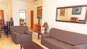 A living room with a sofa, armchair, and coffee table. There is a wooden cabinet, a lamp, and a painting on the wall. - Las Olas 206 by PRP (Playa del Carmen)