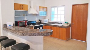 A kitchen with granite countertops, a stove, a microwave, and wooden cabinets. There are three bar stools and a window with blinds. - Las Olas 206 by PRP (Playa del Carmen)