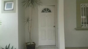 A white door with a small window above it, a potted plant to the left, and a window with decorative bars to the right. - Casa Lupita (Mazatlán)