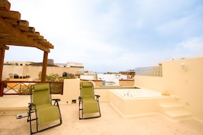 Relax on the rooftop terrace with a hot tub and lounge chairs under a shaded pergola, offering a view of the surrounding buildings.