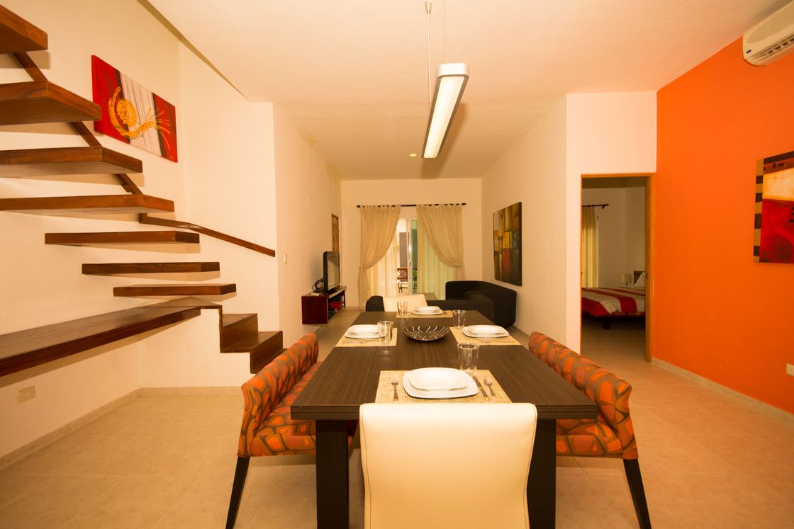 Un coin repas avec une longue table dressée pour quatre personnes, entourée de chaises aux motifs orange. Un escalier avec des marches en bois est visible sur la gauche, et un mur orange se trouve sur la droite.