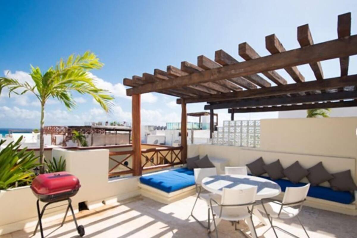 Eine Dachterrasse mit einer Holz-Pergola, einer Sofagruppe, einem runden Tisch und einem Grillwagen.