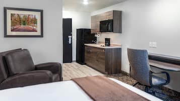A hotel room with a bed, a desk, a chair, a kitchenette, and a framed picture on the wall.