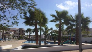 Zona de piscina con palmeras, un tobogán y un edificio al fondo.