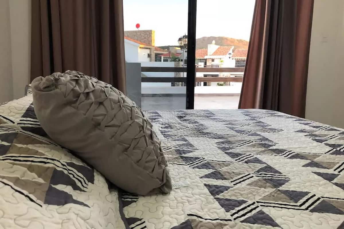 Un lit recouvert d’une courtepointe à motifs et agrémenté d’un coussin décoratif, une fenêtre avec des rideaux bruns, et une vue sur des bâtiments et un ballon rouge au loin.