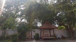 A gazebo with a wooden roof and a bench, surrounded by trees and a concrete ground. - Wanjai Resort (Warin Chamrap)