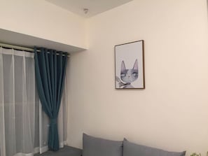 Una habitación con una ventana cubierta por cortinas blancas translúcidas y una cortina azul opaca, un sofá gris y un cuadro con un gato en la pared.